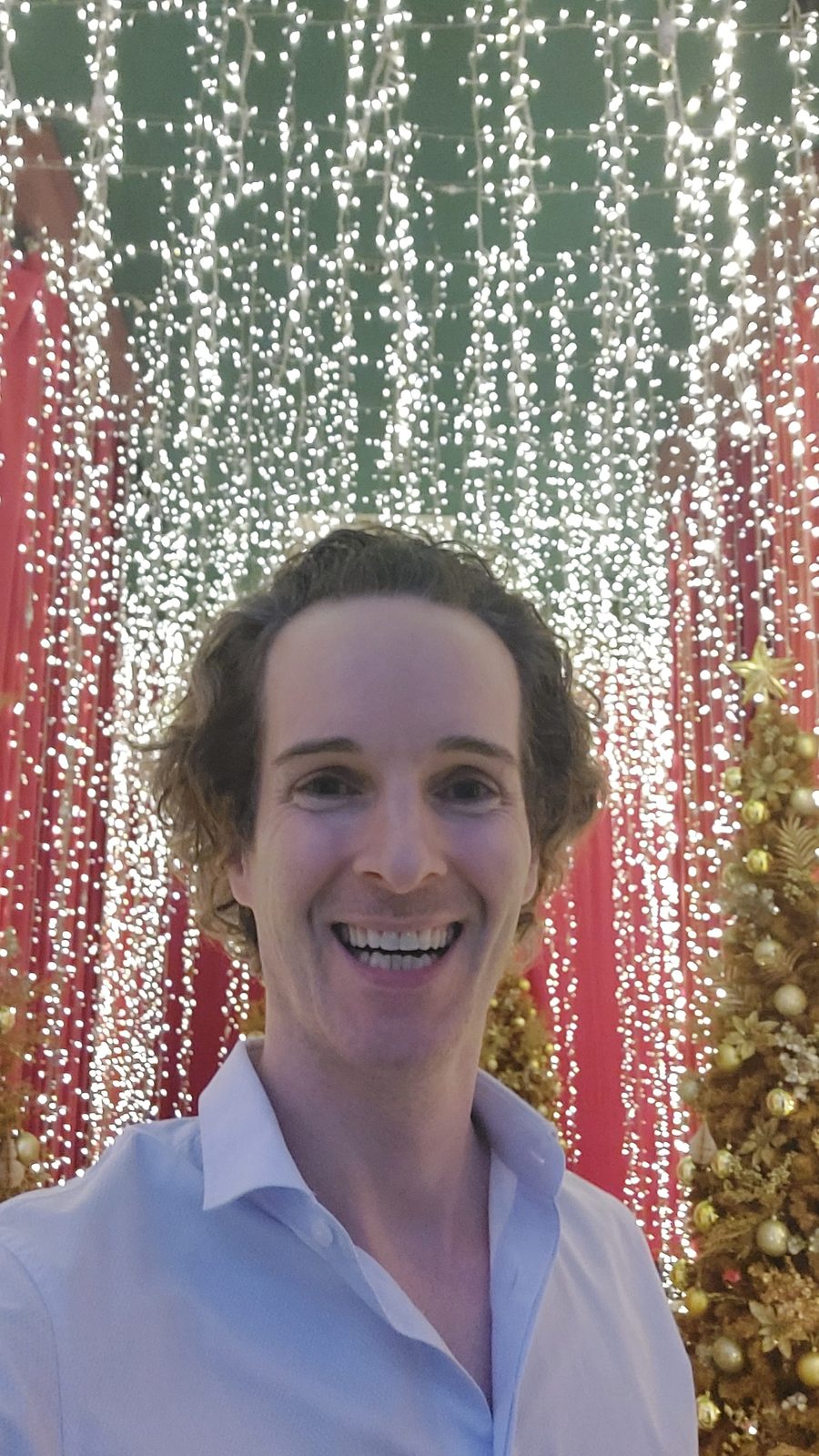 Nikolas Prehn (Felix Nikolas Prehn), in a light blue shirt in front of a wall of fairy lights and a Christmas tree